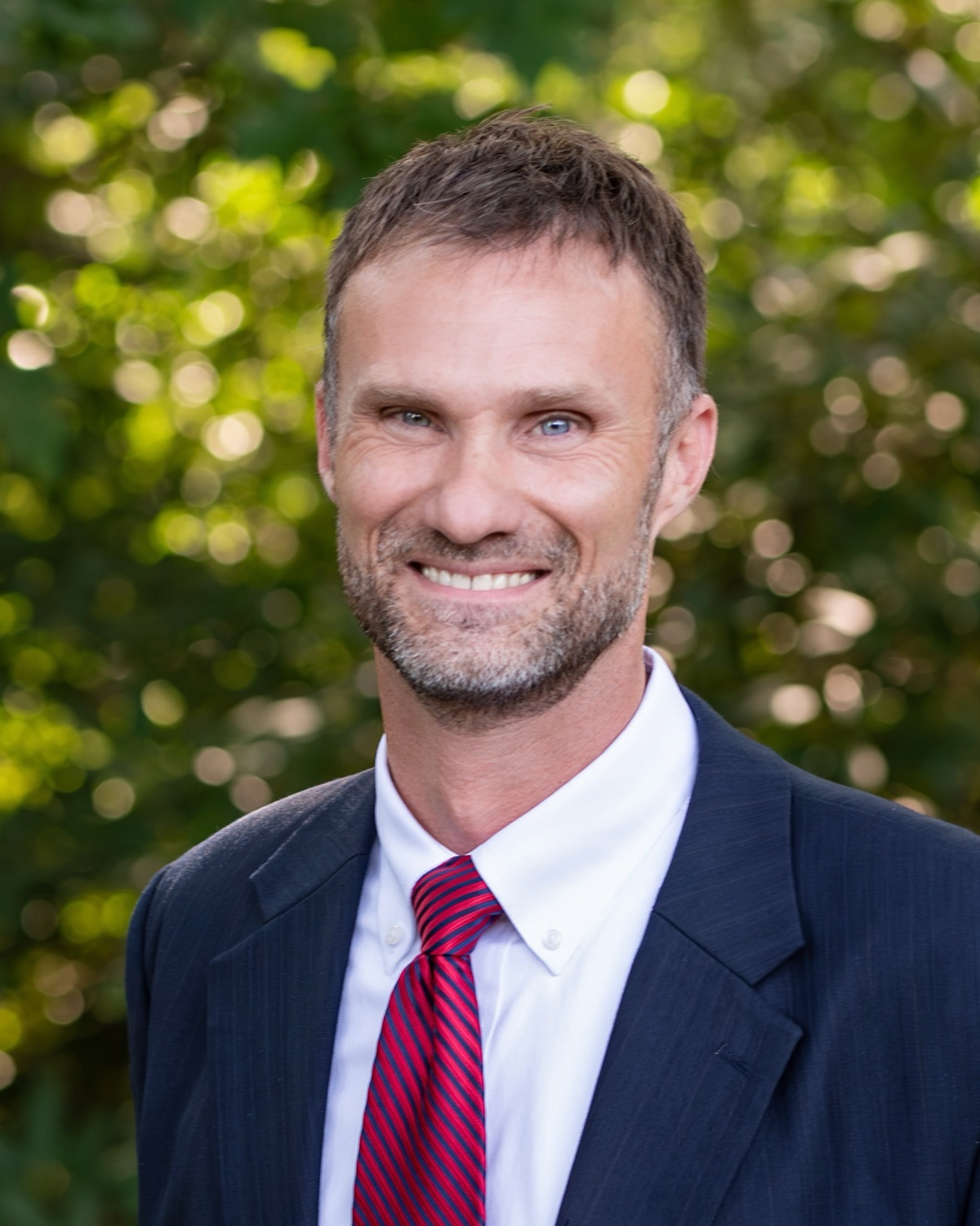 Pastor Ed Kramer, in a navy suit and red tie, in front of summer trees.